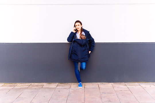 Young Adult Woman Talking On The Smartphone Carrying Her Baby Leaning On A Gray And White Wall With A Serious Gesture Receiving Bad News