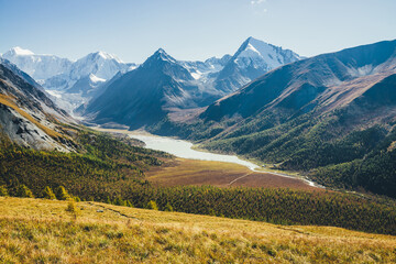 Fototapeta premium Wonderful alpine landscape with mountain lake and mountain river in valley with forest in autumn colors on background of snowy mountains silhouettes under blue sky. Beautiful mountain valley in autumn