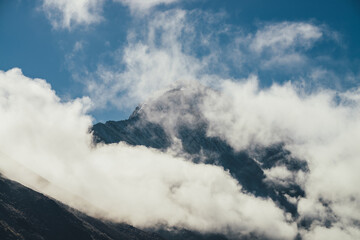 Minimalist view of snow-capped mountain silhouette above thick clouds. Scenic mountain landscape with white-snow sharp peak among dense low clouds in blue sky. Wonderful scenery with snowy pinnacle.