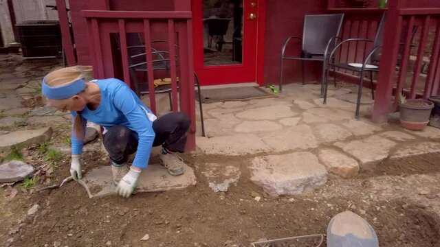 Woman with trowel pressing dirt under rock to set large stone paver into place.