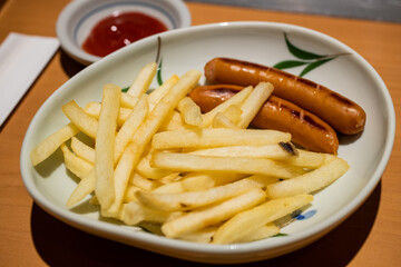 Close-up of french fries with grilled sausage serving as appetiser on the dish dipping with tomato sauce.