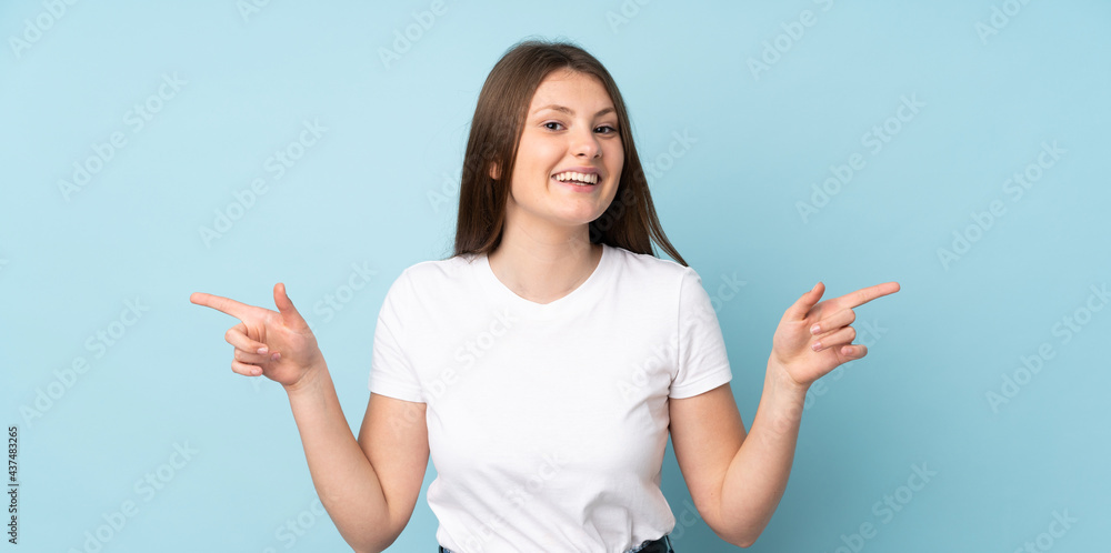 Wall mural teenager caucasian girl isolated on blue background pointing finger to the laterals and happy