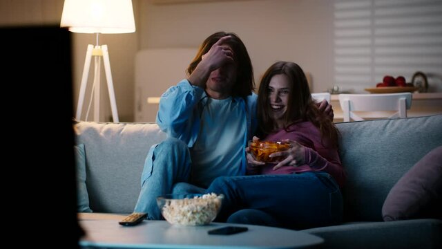 Young man and woman taking chips and popcorn and sitting down on sofa and watching tv at home in evening