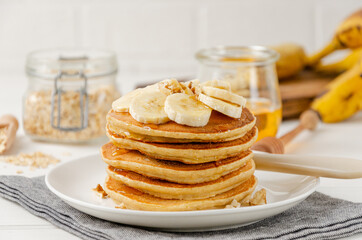 A stack of oatmeal banana pancakes with slices of fresh bananas, walnuts and honey on top with cup of tea on a white wooden background. A healthy breakfast. Copy space.