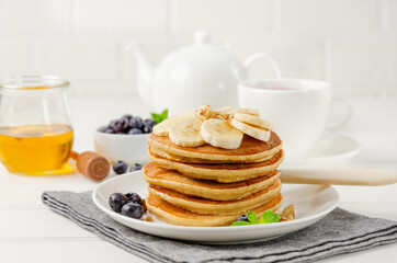 A stack of oatmeal banana pancakes with slices of fresh bananas, walnuts and honey on top with cup of tea on a white wooden background. A healthy breakfast. Copy space.