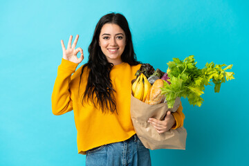Young woman holding a grocery shopping bag isolated on blue background showing ok sign with fingers
