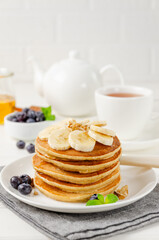 A stack of oatmeal banana pancakes with slices of fresh bananas, walnuts and honey on top with cup of tea on a white wooden background. A healthy breakfast. Copy space.