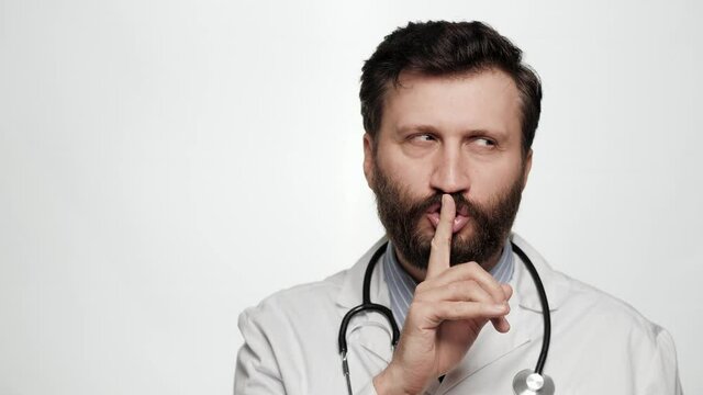 Shh, doctor secret finger. Suspicious smiling man doctor on white background looking at camera and brings his index finger to his mouth lips and she say shhh