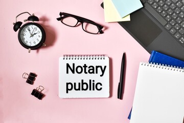 NOTARY PUBLIC is written in a notepad near a laptop, alarm clock, paper and a pen on a pink...