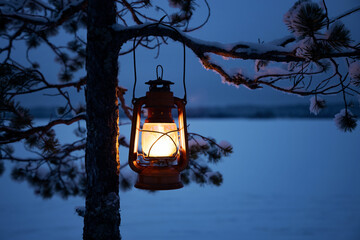  Vintage lantern hanging on snow covered pine tree at night. Oil lamp burning bright. Ice covered lake on background. Finnish nature. 