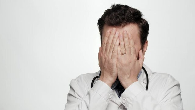 Doctor FACEPALM. Astonished man doctor on white background looking at camera and covers his face with his hands expressing bewilderment