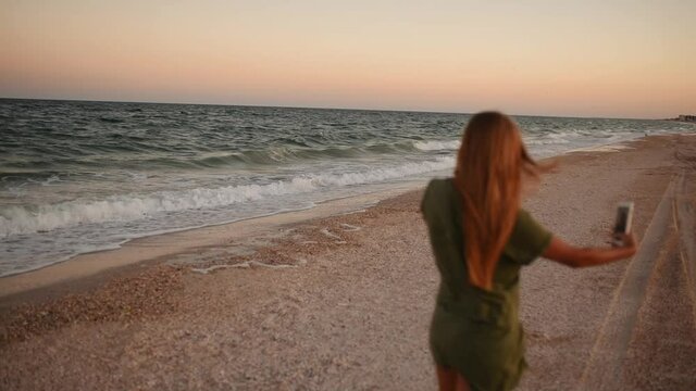 young blonde woman makes selfie after sunset on the beach near the sea. Woman take selfie on smartphone in front of sunrise over the ocean. High quality FullHD footage