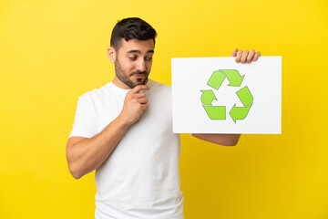 Young handsome caucasian man isolated on yellow background holding a placard with recycle icon