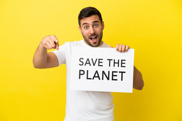 Young handsome caucasian man isolated on yellow background holding a placard with text Save the Planet and pointing to the front