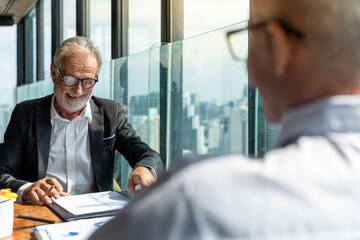 Picture of two business man discussing business and seem like an agreement is made. The young man is wearing white shirt with black tie. The older man is wearing black suit. They meet in hotel lobby.