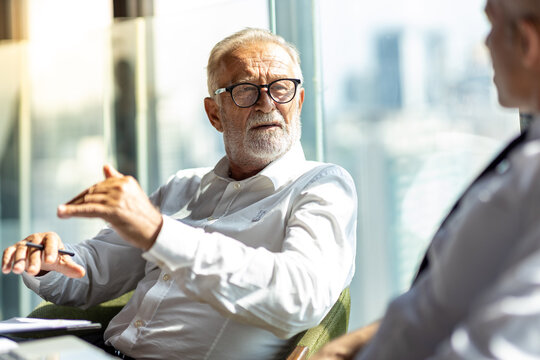 Picture Of Two Business Man Discussing Business And Seem Like An Agreement Is Made. The Young Man Is Wearing White Shirt With Black Tie. The Older Man Is Wearing Black Suit. They Meet In Hotel Lobby.