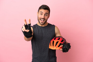 Young cyclist man isolated on pink background smiling and showing victory sign - obrazy, fototapety, plakaty