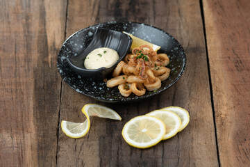 A plate of crunchy deep fried squid rings, calamari with  smoky red bell pepper, almond, cherry vinegar, tomato and cayenne served with tartar sauce dip on the side isolated on wooden table