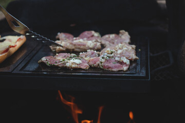 Man uses a steel ladle to turn a loaded pork neck with basil, salt, pepper and oil from one seared side to the other. Barbecue on a fierce outdoor fire. Barbecue season has begun