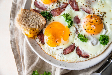Fried eggs with sausage and parsley on a ceramic plate served with bread, light concrete background. Classic breakfast.