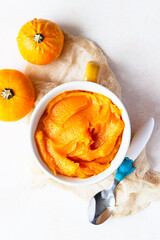 Homemade pumpkin puree in bowl with baby spoon and fresh pumpkins on light concrete background. The...
