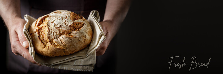 Male hands with fresh baked rye wheat bread on a cotton towel. Dark bakery background banner with...