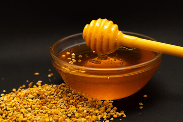 A yellow spoon of honey dipped in a jar of honey and bee bread scattered on a black background....