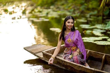 Tuinposter Beautiful girl in purple Thai dress sitting on a wooden boat in the lotus pond. © aTick