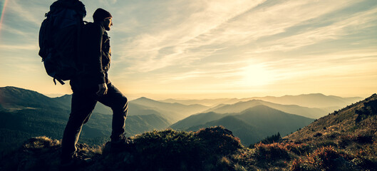 The man standing with a camping backpack on a rock with a picturesque sunset