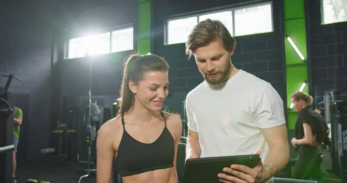 Waist up portrait view of the calm sports girl looking something at the tablet screen with her coach or trainer while making up a training program. Sport and recreation concept