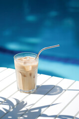 Ice coffee Cyprus Frappe Fredo against blue clear water of the swimming pool, on white table, with metal straw . Summer minimalistic background, holiday or vacation concept. Sun and shadows.Copy space