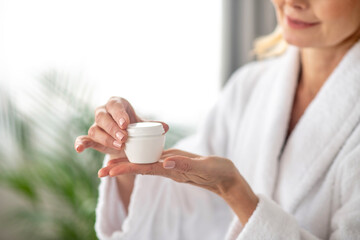 A woman in a white bath robe holding a jar of a facial cream