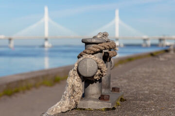 Fototapeta premium Old rope and mooring double cross bollard in sailing port. Cable-stayed bridge and blue water in the background. Summer sea travel concept