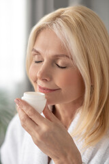 A woman in a white bath robe holding a jar of a facial cream and smelling it