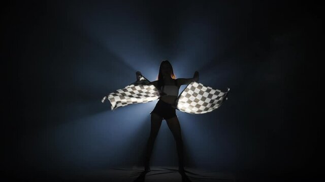 Silhouette of young woman waving a checkered race flag to signal the start of a racing event. Brunette posing full length in a dark smoky studio with backlight. Slow motion ready, 4K at 59.94fps.