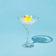 Creative composition with yellow rubber duck and bubble bath foam in martini cocktail glass on pastel blue background. Surreal bathing concept. Retro style aesthetic idea.