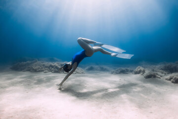Lady freediver with fins posing and glides underwater in blue sea with sunlight.