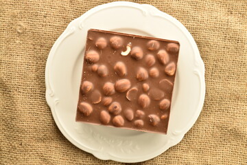 One bar of dark chocolate with nuts on a white saucer on a jute napkin, close-up, top view.