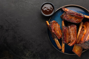 Smoked beef ribs with bone and barbecue sauce on a round dish, top view, rustic black background....
