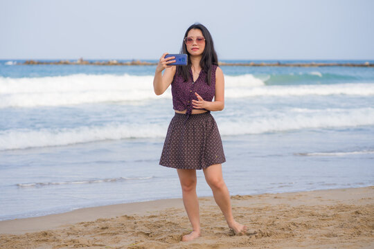 Asian woman taking selfie with mobile phone - lifestyle photo of young happy and beautiful Korean girl taking self portrait on beach smiling cheerful and relaxed enjoying holidays