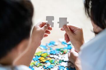 Back view of mother and son lying on the floor holding puzzle pieces and putting them together. Happy family during work at home. Woman teaches child to solve puzzles. Parent and cheerful concept.