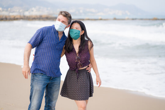 covid19 beach holidays - young beautiful and happy mixed ethnicity couple of Asian woman and Caucasian man in face mask walking playful enjoying love and romantic walk