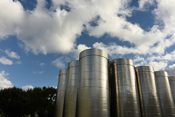 A row of outside stainless steel fermentation wine tanks against clouds and a blue sky
