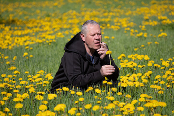 A man, an adult, sits in a dandelion field, holding a cigarette in one hand and a yellow flower in the other. Concept, pondered about the meaning of life, looks at nature