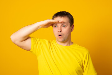 Young blond man wearing casual yellow shirt very happy and smiling looking far away with hand over head. Searching concept.