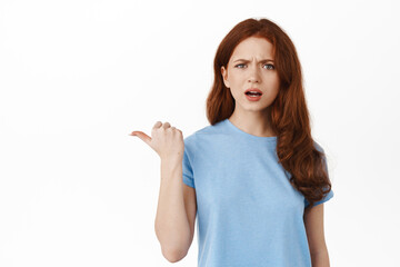 Confused and annoyed redhead girl pointing finger left, showing something strange and frustrating, cant understand what is that aside, standing in t-shirt against white background