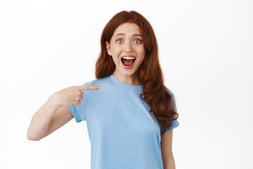 Amazed young natural woman, student with red hair in t-shirt pointing at herself with wondered and surprised face, being chosen, self-promoting, winning unexpectedly, white background