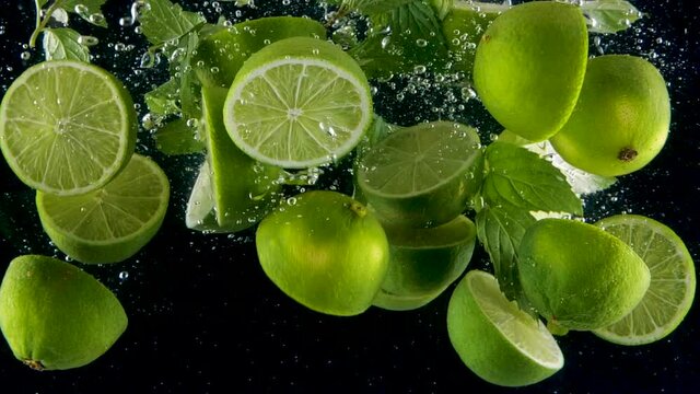 Limes with mint leaves splashing into the water in slow motion. Lots of limes falling slowly on a black background. Organic green citrus isolated on black background. Fresh water with fruits.