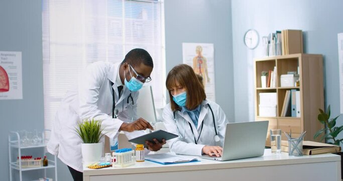 Young African-American intern in protective medical mask consults with female doctor about treating patient by showing results of tests on tablet.