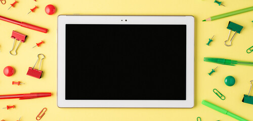 Overhead photo of tablet monochrome green and red stationery pencil pen paperclips and felt-tip isolated on the yellow background
